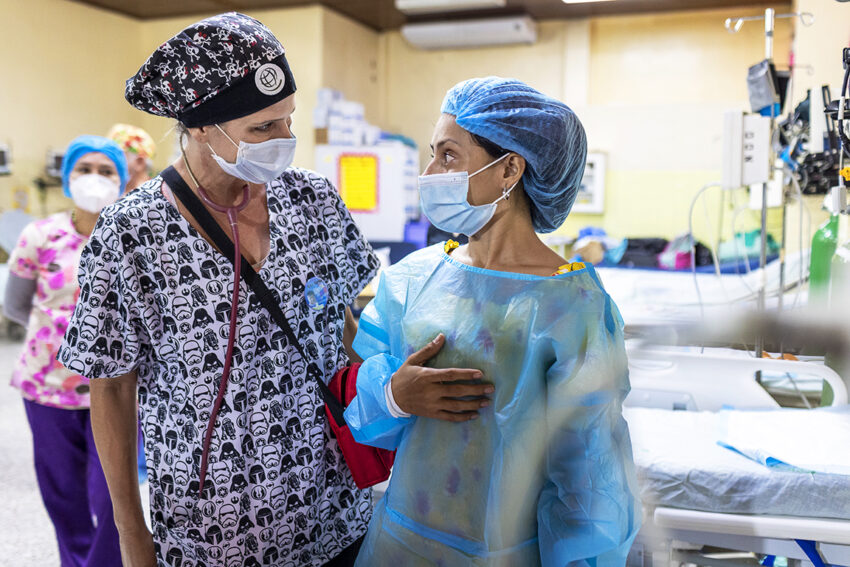 A medical professional wearing surgical scrubs has her arm around a woman waiting by a hospital bed.
