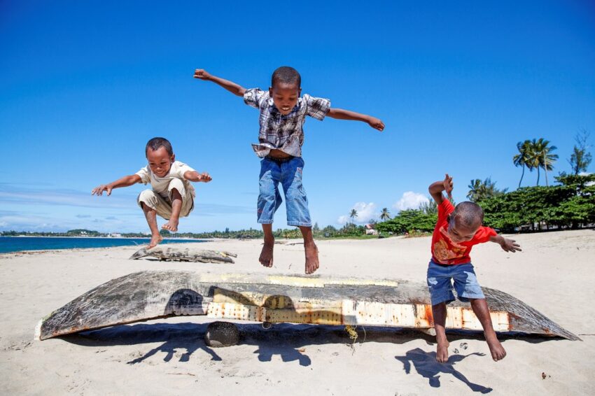 Three young boys playfully jump off an overturned boat on the sand